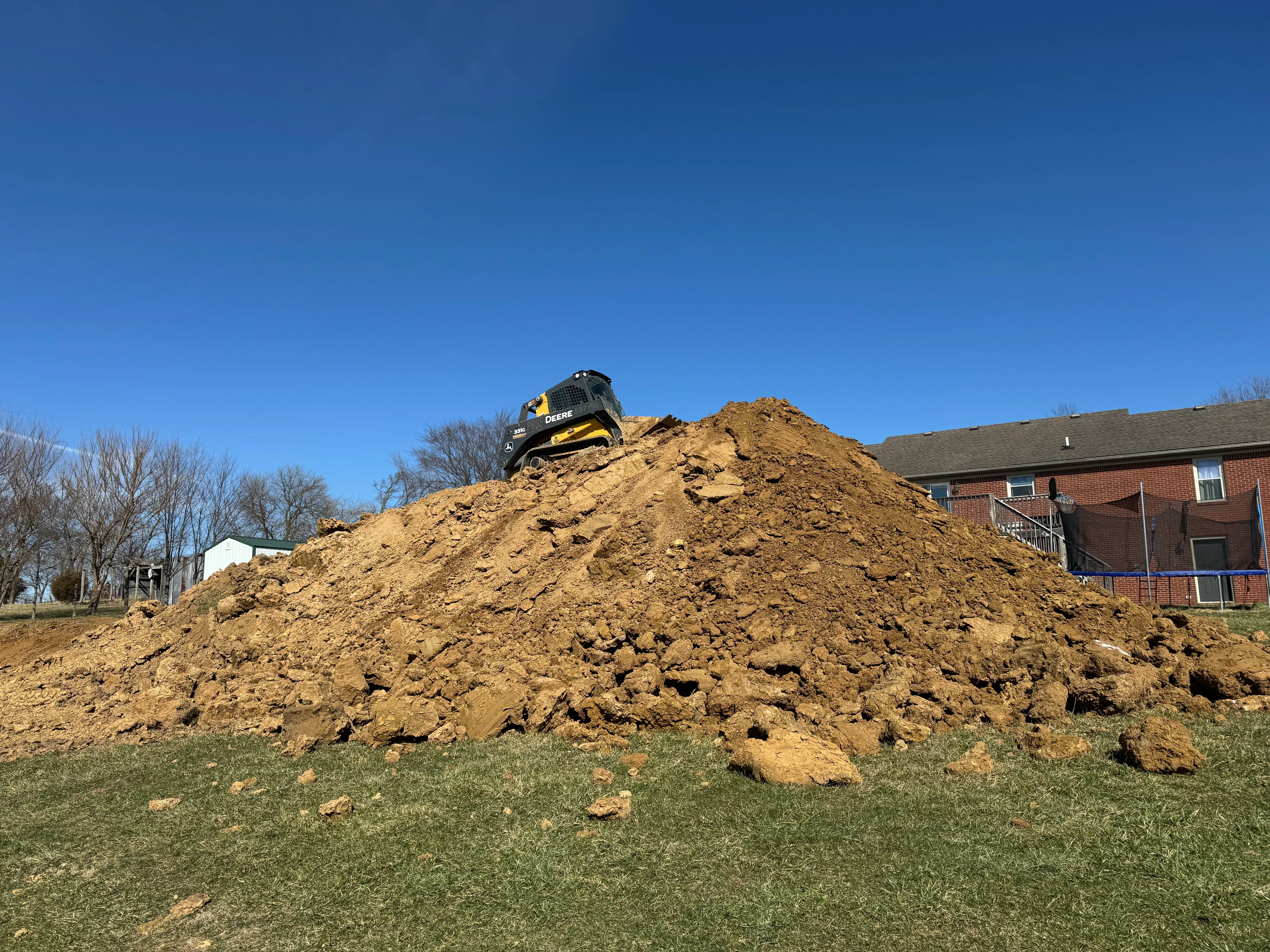 Yellow and black excavator on large soil stockpile with residential brick building in background