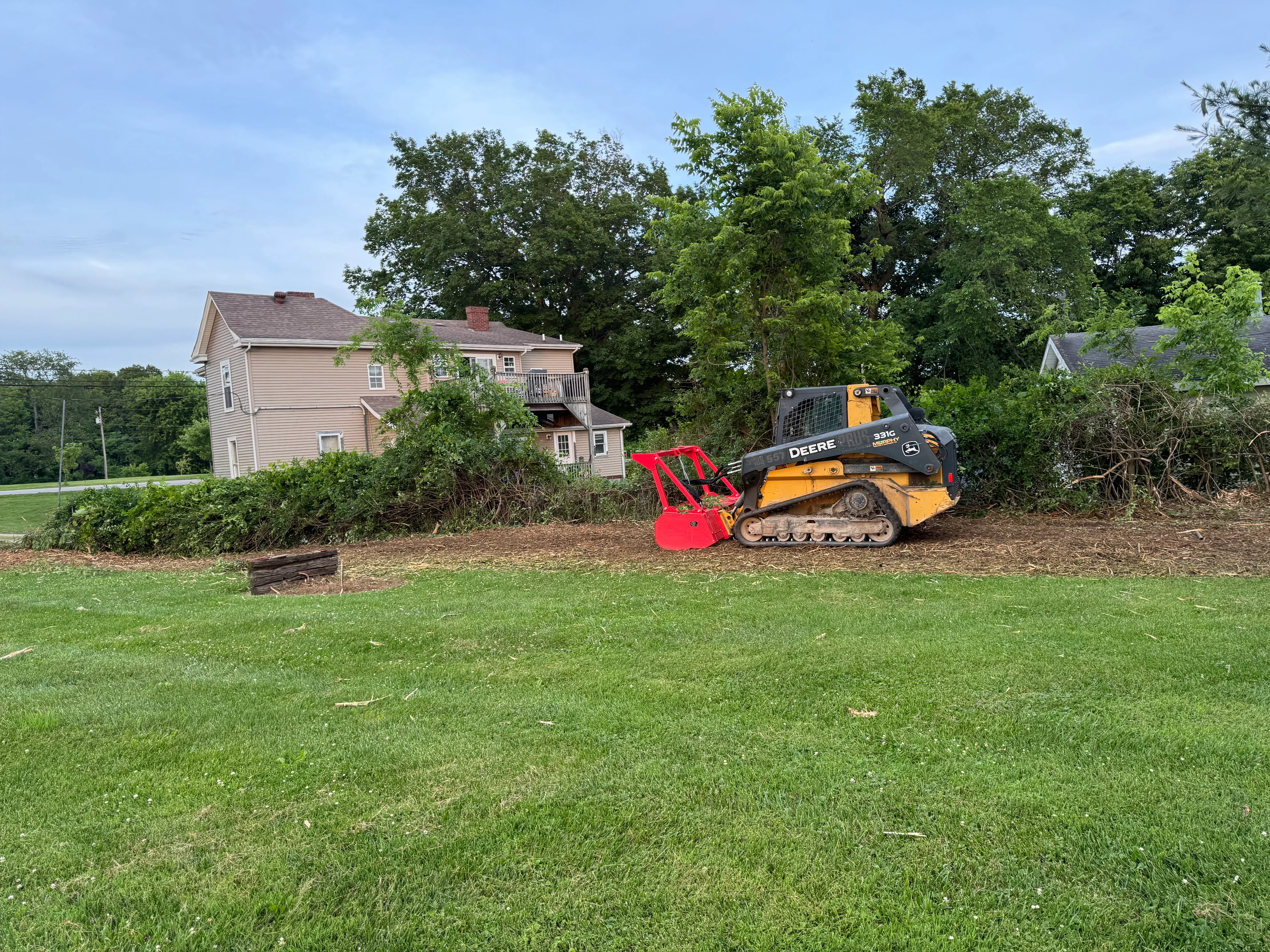 Tracked skid steer with rotary cutter clearing brush and vegetation from residential property
